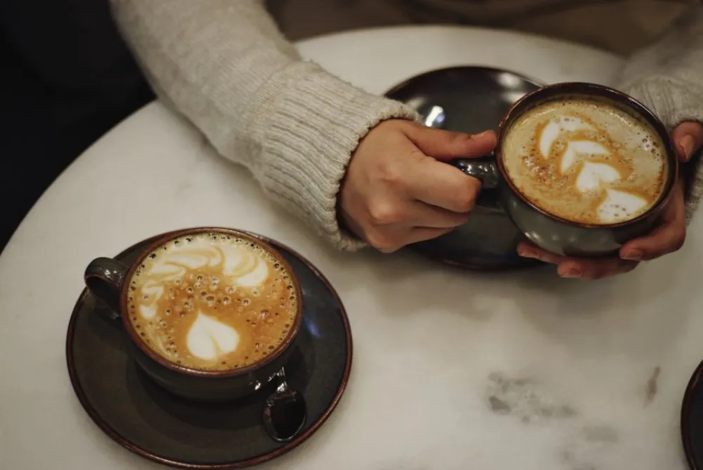 Close-up of two latte art cups on a table held by cozy sweater-clad hands.
