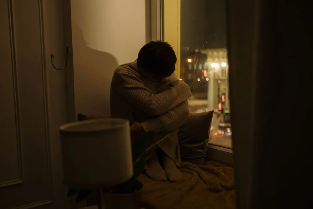 A woman sits alone by a window at night, depicting solitude and reflection.