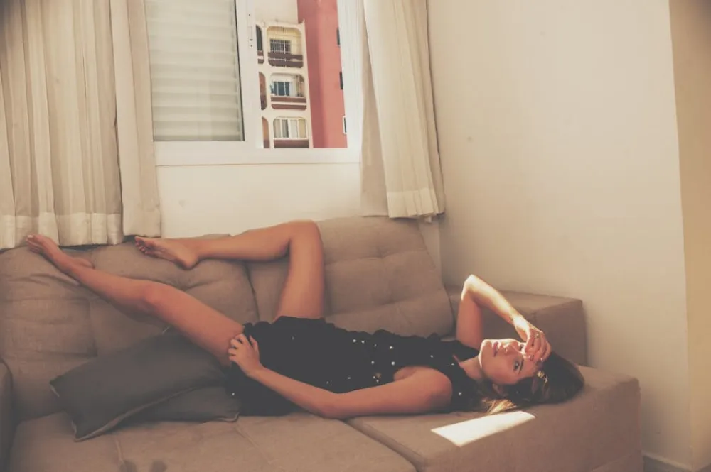 A woman in a black dress lying on a beige couch by a window with sunlight streaming in.