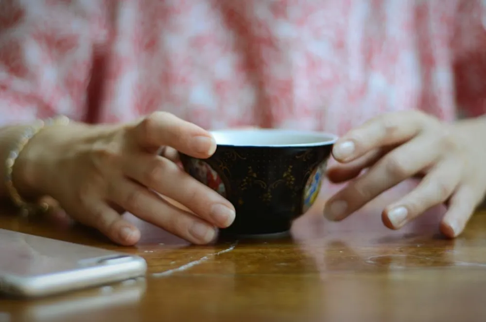 Hands holding a decorative cup on a wooden table, reflecting morning relaxation indoors.
